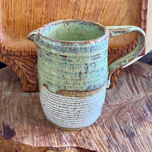 May include: Handmade ceramic pitcher with a light green and brown glaze. The pitcher has a spout and a handle. The bottom half features horizontal textured lines. The pitcher is sitting on a wooden surface.