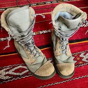 May include: A pair of military-style boots in shades of green and tan, with gray laces. The boots have a suede toe cap and a padded collar. The boots are on a red patterned rug.