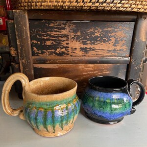 May include: Two handmade ceramic mugs. One mug is tan with green and blue glaze, featuring a large handle. The other mug is black with blue and green glaze. Both mugs are on a light-colored surface.