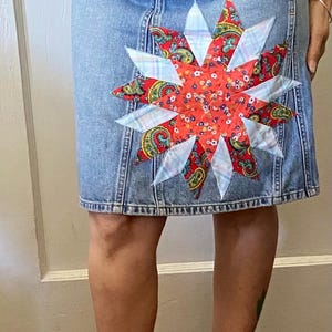 May include: A denim skirt with a large patchwork star design in red, white, and blue fabrics. The skirt is knee-length and has a classic five-pocket style.