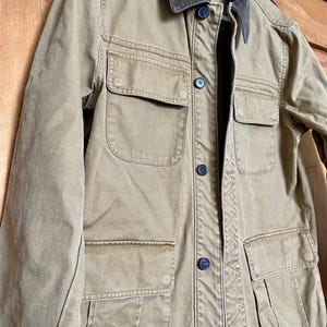 May include: A tan-colored jacket with a corduroy collar and multiple front pockets. The jacket has a button-up closure and a classic, utilitarian design. The jacket is made of a durable fabric and is suitable for various weather conditions.