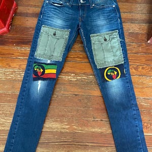 May include: Blue denim jeans with two olive green cargo pockets and patches featuring a Rastafarian flag design. The jeans have a classic five-pocket design and a slim fit.
