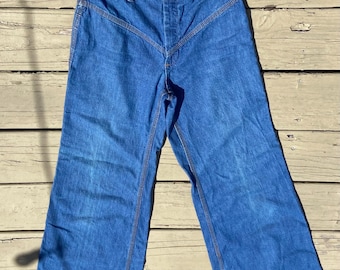 May include: Blue denim pants with a high waist and wide legs. The pants have a button and zipper closure, with a distinctive V-shaped seam detail at the front. The seams are highlighted with a contrasting orange thread.