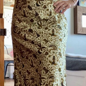 May include: A cream-colored, short-sleeved dress with a brown and green floral pattern. The dress has a V-neckline and a fitted silhouette. The fabric appears to be lightweight, suitable for warm weather. The dress is a maxi length.