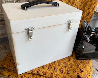 May include: White wooden box with a black handle and silver clasps. The box is sitting on a yellow and red patterned fabric.