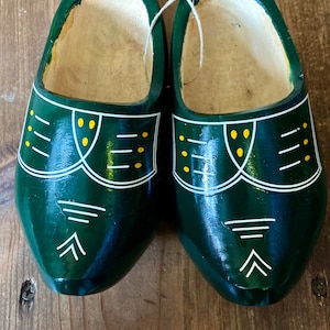 May include: A pair of dark green wooden clogs with white and yellow decorative accents. The clogs have a traditional design, with a rounded toe and a curved shape. They are displayed on a wooden surface.