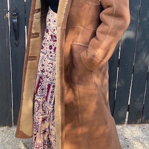 May include: A long brown suede coat with a shearling lining. The coat is worn over a patterned skirt.