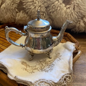 May include: A silver teapot with a lid and a decorative handle. The teapot has three feet and is sitting on a white cloth with lace trim.