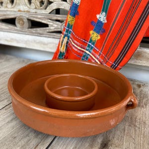 May include: A terracotta-colored ceramic serving dish with a central bowl. The dish has a wide, shallow design with two small handles. A vibrant red textile with colorful embroidery is in the background.