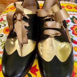 May include: Black leather shoes with gold leather accents and beige satin ribbon laces tied in bows.