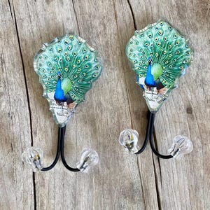 May include: Two decorative wall hooks featuring a peacock design. Each hook has a clear, ornate top with a peacock illustration in shades of blue and green. The hooks have black metal stems and clear, faceted knobs.
