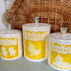 May include: Set of three vintage ceramic canisters with lids. The canisters are off-white with yellow panels featuring the words "TEA", "SUGAR", and "COFFEE". Each canister has a silver-colored knob on the lid and a decorative chicken design.