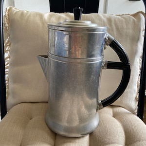 May include: A silver coffee percolator with a black handle and lid. The percolator is sitting on a white, textured surface.