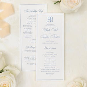 May include: Two rectangular wedding program cards with blue text and a monogram, featuring the names Amelia and Brayden. The cards are surrounded by white roses, a spool of beige silk ribbon, and a small pink velvet ring box with two rings.