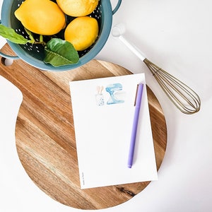 May include: A white notepad with a blue and white illustration of a kitchen mixer and a glass of milk with a straw. A purple pen is on the notepad, which is sitting on a wooden cutting board. A blue colander with lemons and green leaves is in the background.