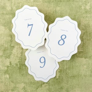 May include: Three white scalloped table number cards featuring the text TABLE NO. and the numbers 7, 8, and 9 printed in a soft blue color. Each card has a thin blue decorative border following the wavy edge of the card stock.