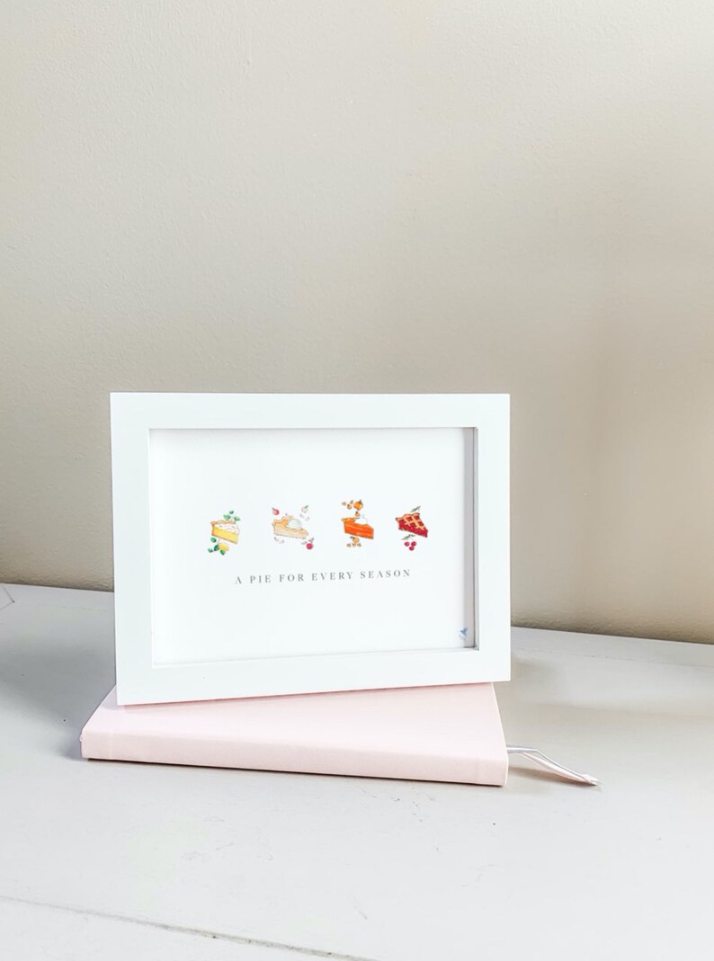 Puede incluir: Una impresi&oacute;n enmarcada en blanco con una ilustraci&oacute;n de acuarela de cuatro tartas diferentes, cada una representando una estaci&oacute;n diferente. El texto "A pie for every season" est&aacute; impreso debajo de las tartas.