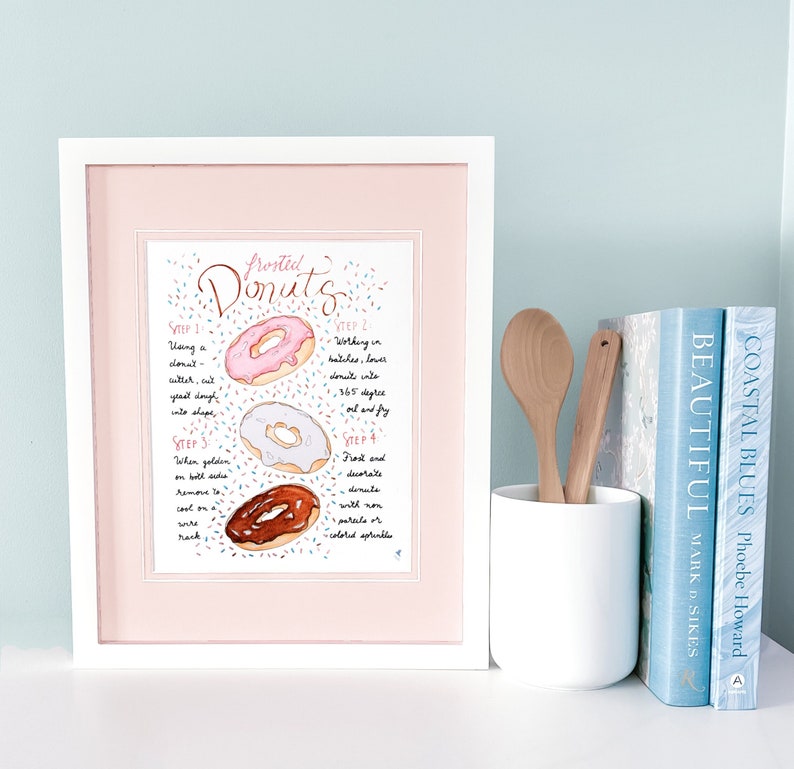 May include: A framed print with a pink background and a white border. The print features a watercolor illustration of three donuts with sprinkles and text that reads "Frosted Donuts" and outlines the steps for making donuts.