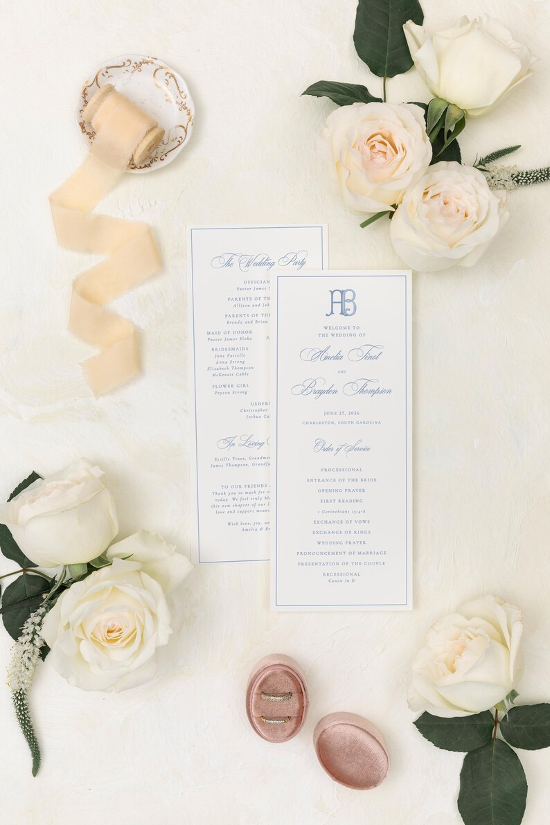 May include: Two rectangular wedding program cards with blue text and a monogram, featuring the names Amelia and Brayden. The cards are surrounded by white roses, a spool of beige silk ribbon, and a small pink velvet ring box with two rings.