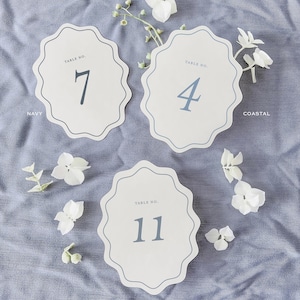 May include: Three scalloped-edge table number cards with navy blue numbers and text. The cards are arranged on a light blue fabric with white floral accents. The cards read "Table No. 7", "Table No. 4", and "Table No. 11".