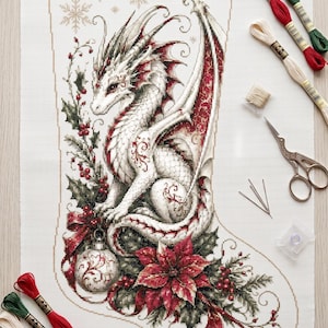 Könnte beinhalten: Ein Kreuzstichmuster auf weißem Stoff mit einem weißen Drachen mit roten Akzenten, umgeben von Stechpalmen, Beeren, einem Weihnachtsstern und einer Christbaumkugel. Enthält Stickgarn in Rot, Grün und Creme, Metallnadeln und eine Schere.