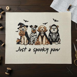May include: A Halloween-themed cross-stitch design featuring four dogs dressed in costumes. One dog wears a witch hat and cape, holding a pumpkin. The text "Just a spooky paw" is below the dogs. Bats are flying above.