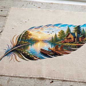 May include: A piece of beige linen fabric featuring a detailed cross-stitch embroidery of a large feather. Inside the feather shape is a landscape scene of a lake, a wooden cabin, a dock with two chairs, a small boat, and a sunset sky.