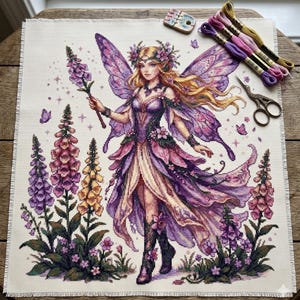 May include: Cross-stitch of a fairy with purple and pink wings, holding a flower wand, surrounded by foxgloves. The fairy wears a floral crown and dress. Embroidery floss, scissors, and a needle minder are next to the cross-stitch.