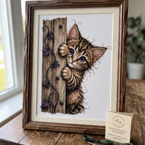 Puede incluir: Una obra de punto de cruz enmarcada titulada The Peekaboo que muestra a un gatito atigrado marrón asomándose detrás de un poste de madera con enredaderas. Incluye una tarjeta informativa, hilo de bordar verde y tijeras doradas.