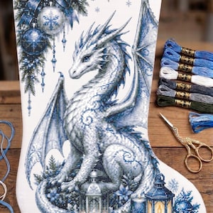 May include: A white fabric Christmas stocking featuring a detailed cross-stitch design of a white dragon, blue ornaments, pine branches, and lanterns. The stocking is accompanied by several skeins of blue and white embroidery thread and gold scissors.