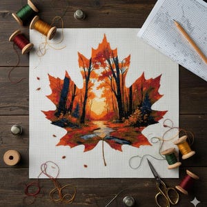 Pode incluir: Um projeto de ponto cruz que retrata uma paisagem de outono dentro de uma folha de bordo. A folha é em tons de laranja e vermelho, com uma cena de floresta no interior. Materiais de costura, incluindo carretéis de linha e dedais, circundam o projeto.