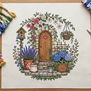 May include: A cross-stitch project on white fabric featuring a stone archway with a wooden door, a heart, a birdhouse, lavender, hydrangeas, and flowers. Includes embroidery floss in various colors, a needle holder, and gold-toned scissors.
