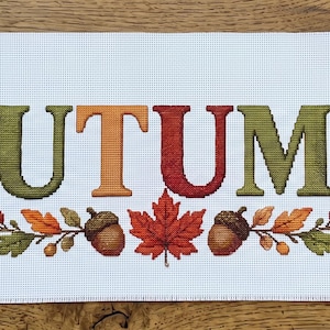 Puede incluir: Un camino de mesa rectangular de tela blanca con la palabra AUTUMN en letras mayúsculas grandes y multicolores. El diseño está flanqueado por calabazas naranjas y blancas, bellotas marrones y hojas de arce rojas, naranjas y verdes.