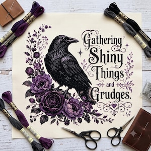 Puede incluir: Un patrón de punto de cruz sobre tela crema con un cuervo negro posado entre rosas y follaje morados. El texto dice "Gathering Shiny Things and Grudges". El diseño está rodeado de hilo de bordar en tonos morado, negro y gris, con dos pares de tijeras y dos estuches para agujas.