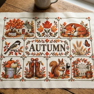 May include: A cross-stitch sampler on cream fabric featuring the word AUTUMN in the center, surrounded by seasonal motifs including a house, a sleeping fox, a bird, wheat, a butterfly, pumpkins in a bucket, boots, a squirrel, and a gate.