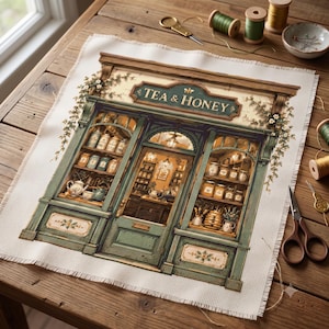 Könnte beinhalten: Eine Kreuzstichstickerei, die ein Ladenlokal mit dem Text TEA & HONEY darstellt. Das Design zeigt eine grüne Holzfassade, Regale voller Gläser und Teekannen sowie Kletterpflanzen. Der Stoff hat rohe, ausgefranste Kanten und liegt auf einem Holztisch.
