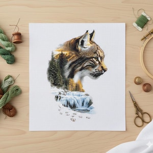 Op de afbeelding: Kruissteekborduurwerk van een lynx in profiel, met gedetailleerde bruine en bruingele vachtpatronen. Het ontwerp bevat een bos- en watervalscène. Ambachtelijke benodigdheden, waaronder garen, schaar en een borduurring, zijn rond het afgewerkte stuk geplaatst.