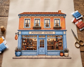 May include: A cross-stitch embroidery on beige fabric depicting a two-story pottery studio with an orange brick facade, blue trim, and a sign reading POTTERY STUDIO. The scene includes shelves of ceramics, a central door, and potted plants.
