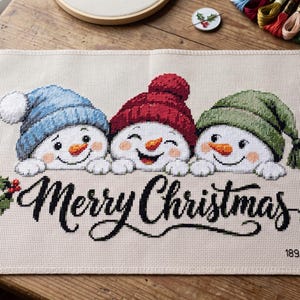 May include: A rectangular cross-stitch fabric piece featuring three snowmen wearing blue, red, and green knit hats. The snowmen have orange carrot noses and are positioned above the black embroidered text Merry Christmas. Holly berries are visible.
