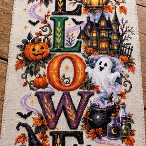 Può includere: Un ricamo a punto croce verticale su tessuto chiaro che riporta la parola HALLOWEEN in grandi lettere colorate. Il disegno include un gatto nero, una casa stregata, un fantasma, un calderone, pozioni, un teschio, libri e zucche autunnali.