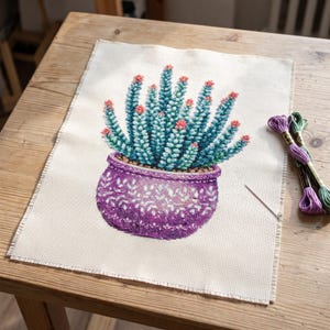 May include: A cross-stitch embroidery project on cream-coloured Aida fabric featuring a teal cactus with pink tips in a purple patterned pot. Beside the fabric are two skeins of embroidery floss in purple and green and a single silver needle.