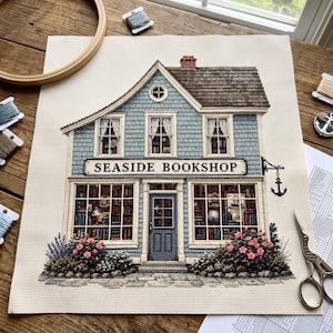 Peut inclure: Un projet de broderie au point de croix sur toile Aida blanche représentant un bâtiment bleu à deux étages avec un toit marron et le texte SEASIDE BOOKSHOP. Le motif présente de grandes vitrines remplies de livres, une porte et des fleurs roses.