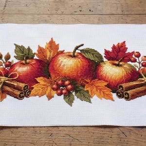 Op de afbeelding: Een rechthoekige witte tafelloper met een centrale horizontale print van vier rode appels, bundels bruine kaneelstokjes vastgebonden met touw, trossen kleine rode bessen en levendige oranje, gele en groene herfstachtige esdoornbladeren.