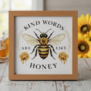Peut inclure: Broderie au point de croix encadrée avec une abeille et l'inscription "KIND WORDS ARE LIKE HONEY". L'abeille est représentée en jaune et noir. Le cadre est en bois clair, et le fond est blanc. L'œuvre est posée sur une surface en bois.