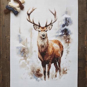 Può includere: Ricamo a punto croce di un maestoso cervo con grandi corna, ambientato in una scena di bosco innevato. L'opera utilizza tonalità di marrone, bianco e blu. Filo da ricamo e forbici visibili.