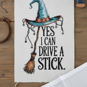 Op de afbeelding: Kruissteekproject met een heksenhoed en bezem, met de tekst "YES I CAN DRIVE A STICK." De hoed is blauwgroen met een bruine band en gesp. Inclusief schaar, draad en borduurring.