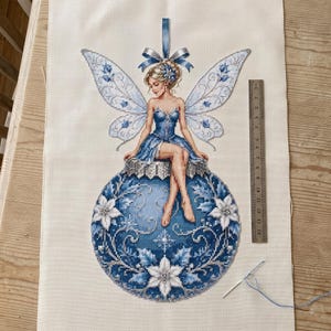 Peut inclure: Une broderie au point de croix sur tissu crème représentant une fée aux ailes bleues assise sur une boule de Noël bleue. Une règle métallique indique que le motif mesure environ 14 cm de large. Une aiguille avec du fil bleu est en bas.
