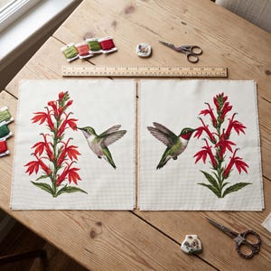 May include: Two square fabric panels featuring cross-stitch designs of hummingbirds hovering near red cardinal flowers. A wooden ruler measuring 18 inches is placed above the panels, accompanied by embroidery floss, scissors, and a needle minder.