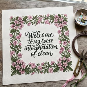 Könnte beinhalten: Ein quadratisches Stück weißer Stoff mit einem Kreuzstich-Design, das einen floralen Rand aus rosa Blüten und grünen Blättern zeigt. Der mittlere Text lautet "Welcome to my loose interpretation of clean" in schwarzer Schreibschrift.