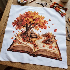 Könnte beinhalten: Ein weißes Stoffquadrat mit einer detaillierten Kreuzstichstickerei eines aufgeschlagenen Buches. Ein großer Baum mit leuchtend orangefarbenen und gelben Herbstblättern wächst aus den Seiten, mit einer kleinen Bank und einem gewundenen Pfad.